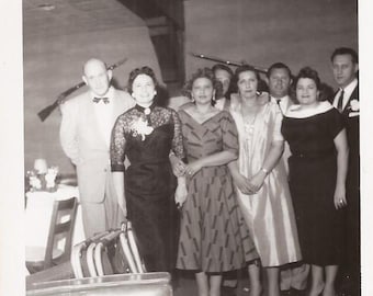 May include: A group of people dressed in formal attire pose for a photo. The women are wearing dresses and the men are wearing suits. The women are smiling and the men are looking at the camera. The photo is taken in a dimly lit room.