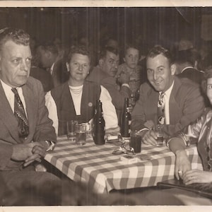 May include: A group of people gathered around a table, some holding drinks. The table is covered with a checkered tablecloth. There are bottles of beer on the table. The people are smiling and appear to be enjoying themselves.