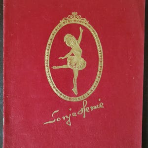 Könnte beinhalten: Ein roter Buchumschlag mit einem goldenen ovalen Rahmen, der eine Ballerina-Illustration zeigt. Die Ballerina trägt einen Tutu und befindet sich in einer Tanzpose. Der Text "Sonja Henie" ist darunter in Gold geschrieben.
