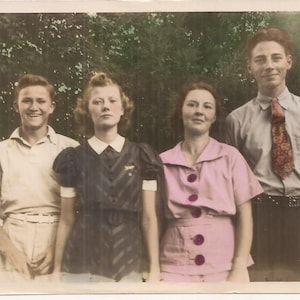 May include: A group of four people standing in front of a green background. Two women are wearing dresses, one man is wearing a white shirt and khaki pants, and another man is wearing a light blue shirt with a patterned tie.