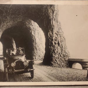 May include: Vintage black and white photograph of a classic car driving through a rock tunnel. The tunnel is carved into a cliffside, with a bridge visible in the distance. The car has a vintage design.