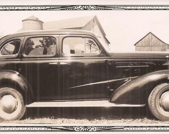May include: Sepia-toned photograph of a vintage black car with a streamlined design. The car has a long hood, curved fenders, and whitewall tires. A barn and silo are visible in the background, framed by a decorative border.