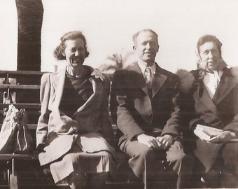 May include: Three people are sitting on a bench outdoors. The person on the left is wearing a coat and a dress. The person in the middle is wearing a suit and tie. The person on the right is wearing a coat and a scarf.