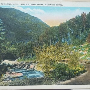 May include: A vintage postcard showing a scenic view of a winding road through a lush green forest. The text on the postcard reads "Nearing Charlemont, Cold River South Turn, Mohawk Trail."