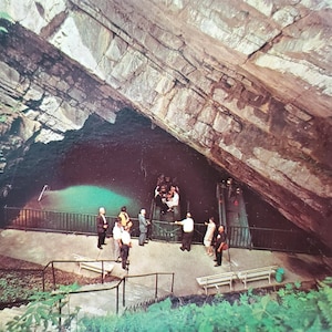 May include: A group of people stand on a platform overlooking a large cave opening. The cave is dark and the opening is filled with a bright blue pool of water. The cave ceiling is made of layered rock.