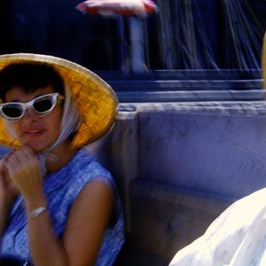 May include: A woman wearing a straw hat and white sunglasses, with a blue dress. The hat is wide-brimmed and yellow. The woman is sitting outdoors, with a second person wearing a similar hat in the background.