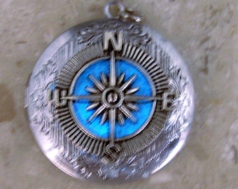 May include: A round silver pendant featuring a central compass rose design set against a vibrant blue enamel background. The compass points are marked with the letters N, S, E, and W. The outer edge of the pendant has an intricate floral engraving.
