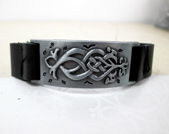 May include: A black leather bracelet with a silver-colored metal centerpiece. The centerpiece features a Celtic knot design with leaf and bird accents. The bracelet is a cuff style.