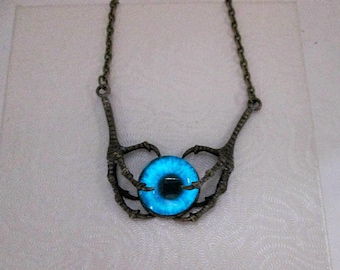 May include: A necklace with a blue eye pendant surrounded by two bronze claw-like hands.