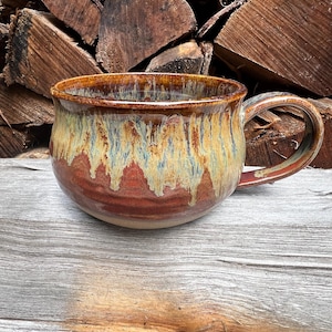 May include: A wide, round ceramic mug featuring a reactive glaze with a deep red base color that transitions into a textured, drippy cream and blue-toned finish. The mug has a sturdy, curved handle and a smooth, rounded rim for drinking.