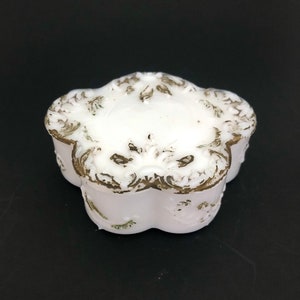 May include: A white porcelain trinket box with a floral design. The box has a raised, ornate design around the top edge and a more subtle floral design on the bottom.