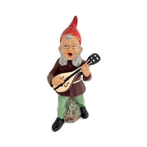 May include: A ceramic gnome figurine playing a lute. The gnome is wearing a red hat, brown tunic, and green pants. The lute is brown with a white and gold design.
