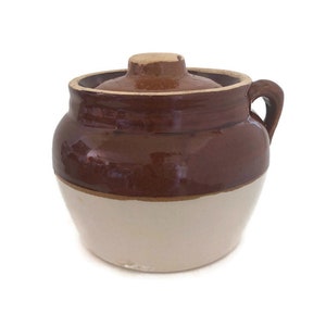 May include: A two-tone ceramic jar with a lid. The upper portion is a glossy brown, while the lower half is a creamy white. It has a small handle and a rounded lid with a knob. The jar is likely used for storage or decoration.