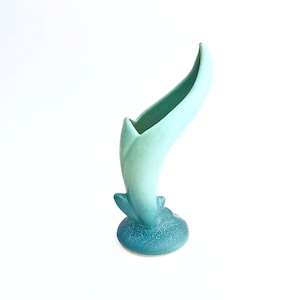 May include: A light blue ceramic vase with a unique, curved design. The vase has a textured base and a smooth, flowing body.