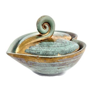 May include: A ceramic bowl with a lid, featuring a teal and gold color scheme. The lid has a spiral handle. The bowl's exterior has a textured, striped pattern. The design is artistic and decorative.