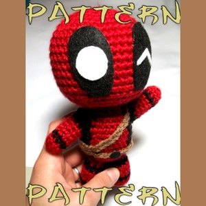 Könnte beinhalten: Häkelmuster für eine rote und schwarze Deadpool Amigurumi-Puppe. Die Puppe hält ihren rechten Arm zum Gruß nach oben. Die Puppe hat eine schwarze Augenmaske mit einem weißen Auge und einem schwarzen Mund.