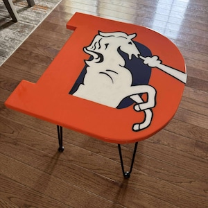 Peut inclure: Table d'appoint orange en forme de lettre "D", ornée d'un logo de cheval blanc et bleu marine. Elle possède des pieds épingles noirs et repose sur un plancher en bois. C'est un élément décoratif.