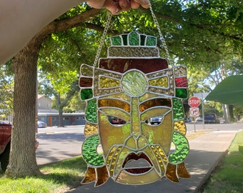 May include: A stained glass mask with a gold, red, and green design. The mask has a large, round face with a mustache and a wide mouth. The mask is hanging from a silver chain.