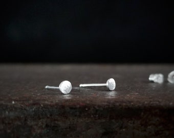 May include: A pair of small, silver stud earrings with a textured, circular design. The earrings are on a dark, textured surface.