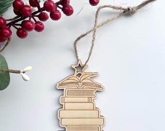 May include: A wooden Christmas ornament shaped like a stack of books, with an open book and star on top. The ornament is made of light-colored wood and has a twine loop for hanging. Red berries and greenery are in the background.
