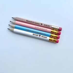 May include: Four wooden pencils with pink, blue, and white paint. The pencils are engraved with the text "Mitchell & Lauren 5.12.24 JACOB AND HAYLEY CHARITY GOLF TOURNAMENT 2024 Bryan Mollie".