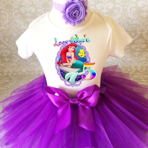 May include: A white bodysuit with a colorful graphic of Ariel from The Little Mermaid, a purple tutu skirt, and a matching headband with a flower. The outfit is designed for a five-year-old's birthday celebration.