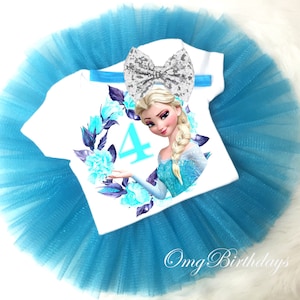 May include: A white t-shirt with a blue floral wreath and a picture of Elsa from Frozen. The number 4 is in the center of the wreath. A silver glitter bow is on top of the shirt. A blue tulle tutu is underneath the shirt.