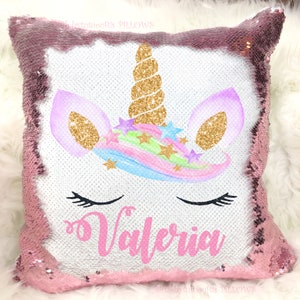 May include: A square pillow with a pink sequin border and a white center. The center features a unicorn design with a gold horn, purple ears, and a rainbow mane. The name "Valeria" is printed in pink.