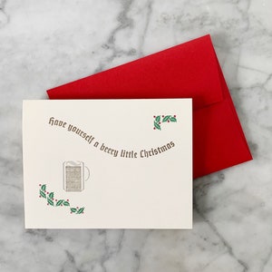 以下が含まれることがあります： 赤い封筒が付いた白いカード。カードには「Have yourself a beery little Christmas」という茶色の文字が書かれている。カードの右上には赤い実のついた緑のヒイラギの枝がある。カードの左には赤い実のついた緑のヒイラギの枝が付いたビールジョッキのスタイリッシュな画像がある。