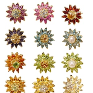 Puede incluir: Doce broches de oro en forma de flor con piedras preciosas de diferentes colores. Cada broche presenta una piedra preciosa central rodeada de piedras preciosas más pequeñas. Las piedras preciosas incluyen amatista, topacio, peridoto, citrino, granate y perlas.