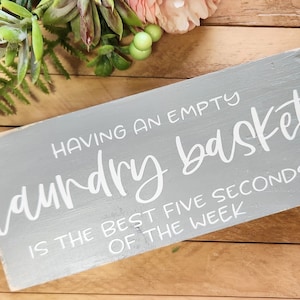 May include: A rectangular gray wooden sign with white painted text that reads: HAVING AN EMPTY laundry basket IS THE BEST FIVE SECONDS OF THE WEEK. The sign has a distressed finish with visible wood grain and slightly worn edges on the corners.