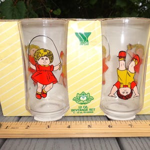 May include: A set of two clear glass tumblers in a yellow cardboard box labeled 12 OZ. BEVERAGE SET. One glass features a child jumping rope, the other shows a child doing a handstand. A wooden ruler shows the box is approximately 8 inches wide.