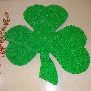 May include: A large, bright green shamrock decoration with a textured surface. The cloverleaf shape is a classic symbol of St. Patrick's Day. The decoration is made of small, textured pieces, and has a hole at the top for hanging.