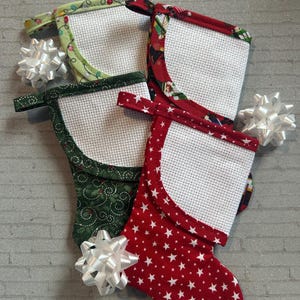 May include: Four small Christmas stockings with white cross-stitch fabric and patterned fabric trim. The stockings have red, green, and light green patterns with white stars, Christmas lights, and holiday designs. White decorative bows are placed near the stockings.