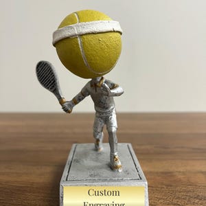 May include: Silver tennis player trophy with a yellow tennis ball head. The base has a gold plaque with the text "Custom Engraving".