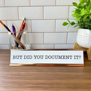 May include: A white desk nameplate with a gold trim and the text "BUT DID YOU DOCUMENT IT?" in black.
