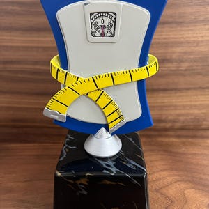 May include: A trophy featuring a blue and white scale design with a yellow measuring tape wrapped around it. The scale reads 0. The trophy sits on a black marble base, symbolizing weight loss or fitness achievements.