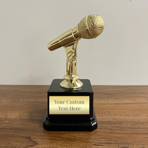 May include: Gold-colored microphone trophy with a black base. The base has a gold-colored plaque with the text "Your Custom Text Here".