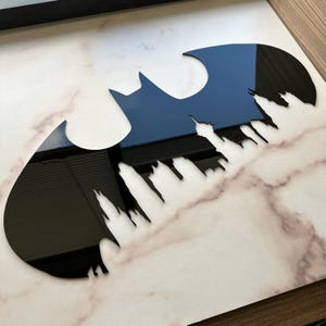 May include: Black Batman logo with a cityscape silhouette cut out. The logo is glossy and reflects the surrounding environment. The background is a marble-patterned surface.