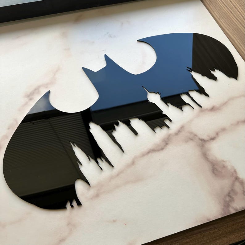 May include: Black Batman logo with a cityscape silhouette cut out. The logo is glossy and reflects the surrounding environment. The background is a marble-patterned surface.