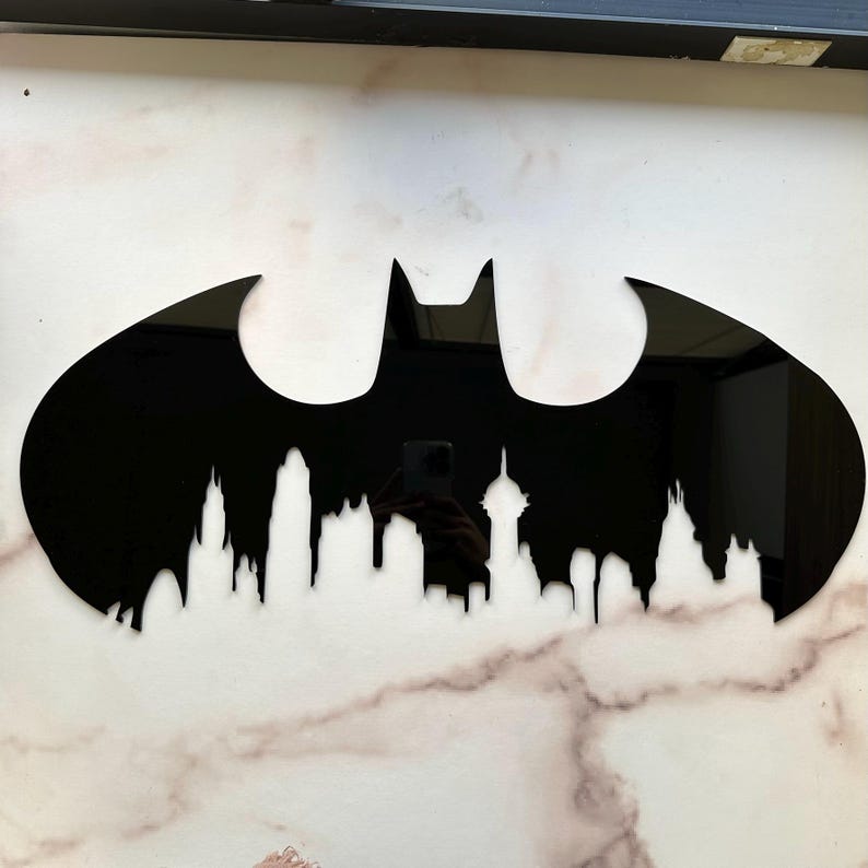 May include: Black Batman logo wall art with a cityscape silhouette at the bottom. The logo is a bat shape with pointed ears and wings. The cityscape features building outlines. The art is mounted on a white and gray marble-patterned surface.