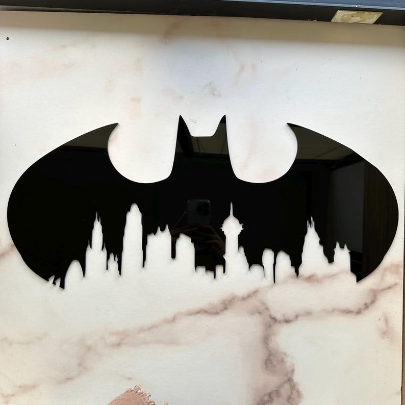 May include: A black Batman logo wall art piece. The logo features a cityscape silhouette at the bottom. The artwork is mounted on a white and gray marble-patterned surface.