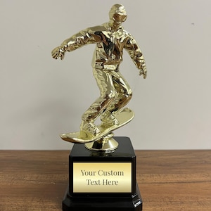 May include: A gold-colored trophy featuring a snowboarder in action, mounted on a black base with a gold plate. The plate reads "Your Custom Text Here". The snowboarder is wearing a jacket, pants, and goggles.