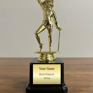 May include: A gold-colored trophy featuring a dancer in a top hat and suit, holding a cane. The trophy is mounted on a black base with a gold plate that reads "Your Name Best Dancer 2024".