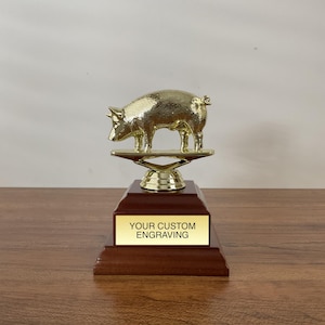 May include: A gold pig trophy with a wooden base. The base has a gold plaque with the text "YOUR CUSTOM ENGRAVING".