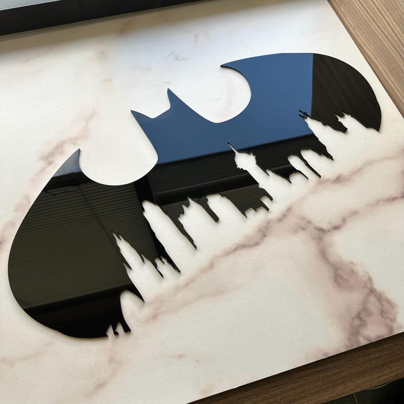 May include: A black acrylic Batman logo with a cityscape silhouette cut out within the emblem. The logo is set against a white and gray marble-patterned surface, creating a striking contrast.