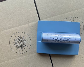May include: A light blue product holder with a clear cylindrical container featuring a black and white floral design. The holder rests on a cardboard box with the text "alkemist organics" and a floral logo.