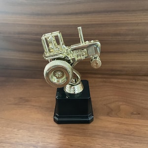 May include: A gold-colored tractor trophy with a black base. The tractor has detailed features, including a large rear tire and a driver's cage. The trophy is displayed on a wooden surface.