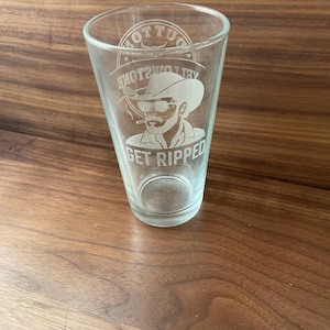 May include: Clear glass pint glass with a cowboy graphic and the words "GET RIPPED". The glass has a flared shape and is suitable for beer or other beverages. The glass is on a wooden surface.