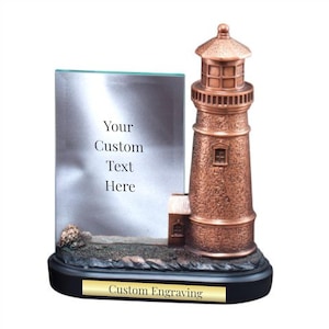 May include: A bronze-colored lighthouse sculpture with a clear glass panel for custom text. The lighthouse has a small building attached and sits on a black base with a gold-colored plate that reads "Custom Engraving".